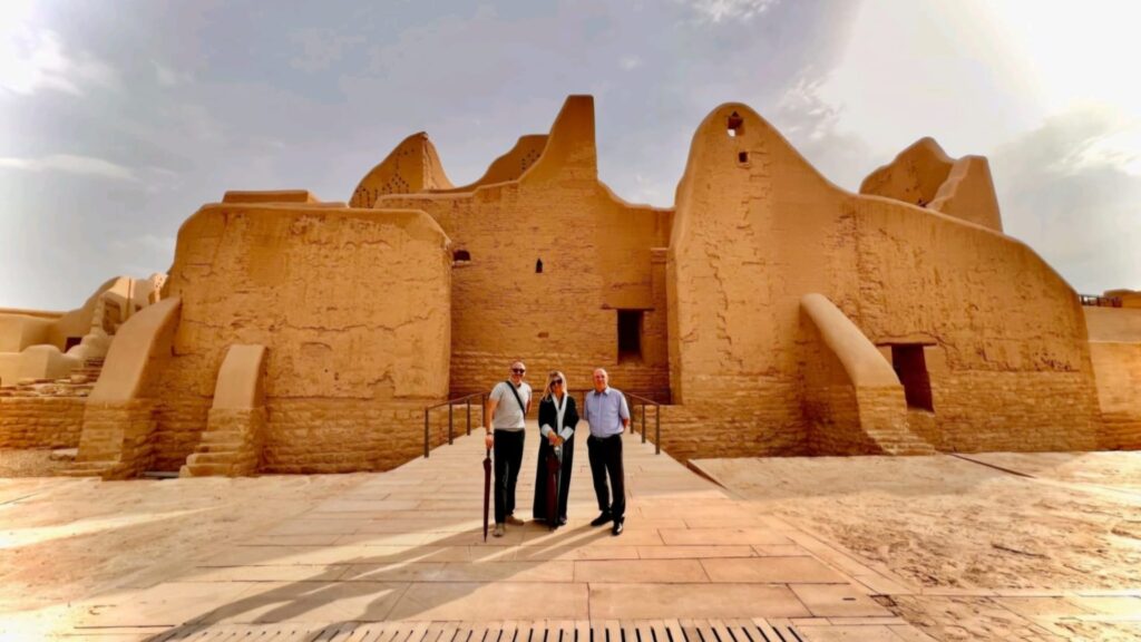 Tre persone visitano un antico edificio in mattoni color sabbia nel deserto sotto cielo nuvoloso.