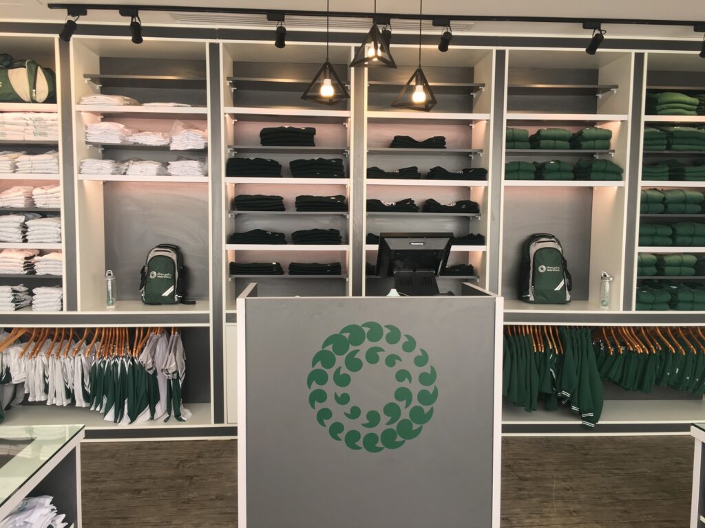 Scaffalatura negozio con asciugamani bianchi e neri, indumenti verdi su grucce, zaini e logo circolare verde